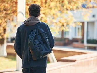 back of young man walking in leafy street wearing backack